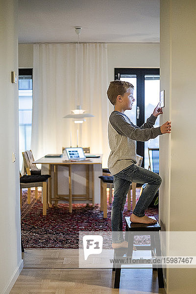 Junge steht auf Hocker  während er ein an der Wand montiertes digitales Tablet im Smart Home benutzt