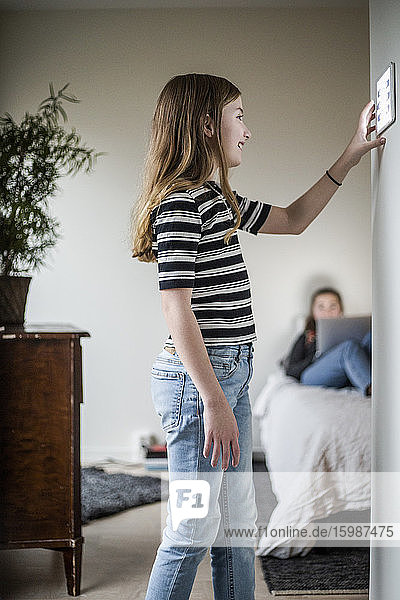 Mädchen benutzt digitales Tablett an der Wand  während die Schwester im Hintergrund im modernen Haus sitzt