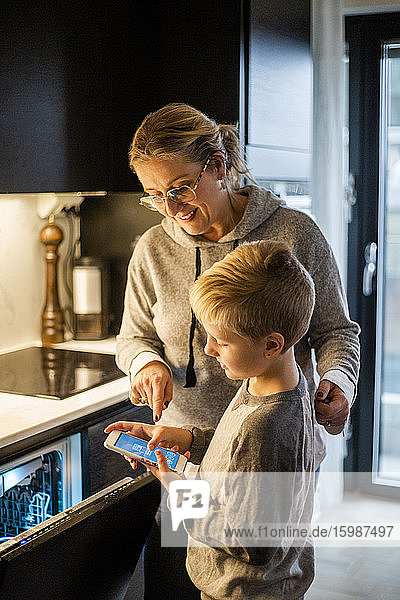 Mutter führt Sohn bei der Benutzung einer mobilen App über den Geschirrspüler in der heimischen Küche