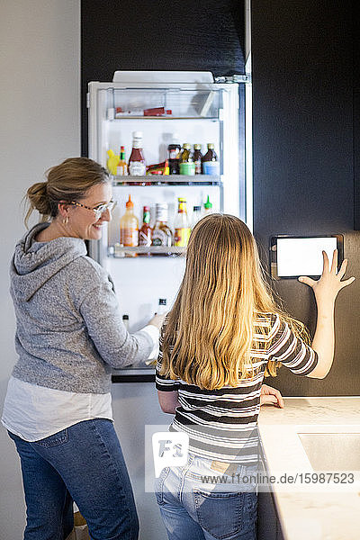 Daughter using digital tablet while mother looking standing by refrigerator at smart home
