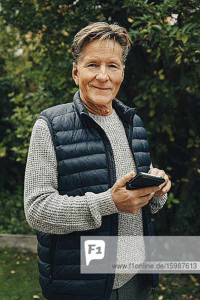 Portrait of smiling senior man holding smart phone while standing at back yard