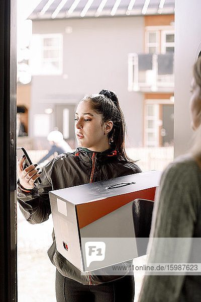 Junge Frau liefert mit dem Mobiltelefon aus  während sie ein Paket zum Kunden bringt