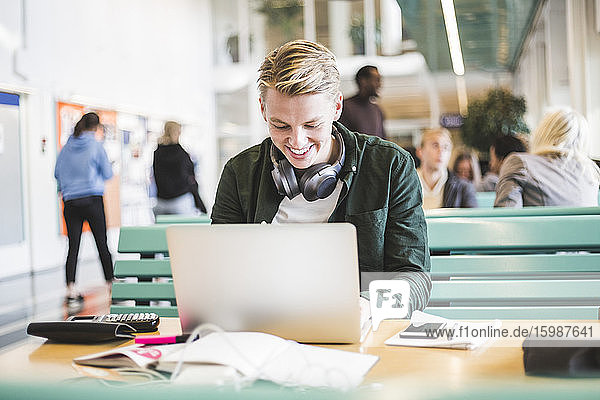 Lächelnder junger männlicher Student benutzt Laptop in der Cafeteria der Universität