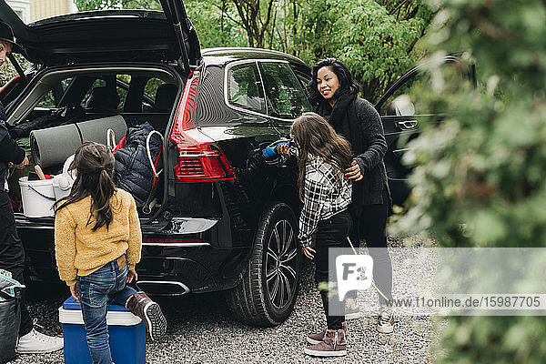 Frau führt Tochter beim Laden eines Elektroautos  während die Familie Gepäck auflädt