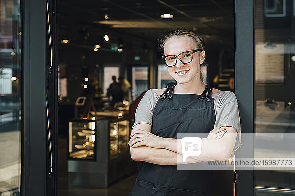 Portrait of smiling andorgynous owner standing with arms crossed at cafe doorway