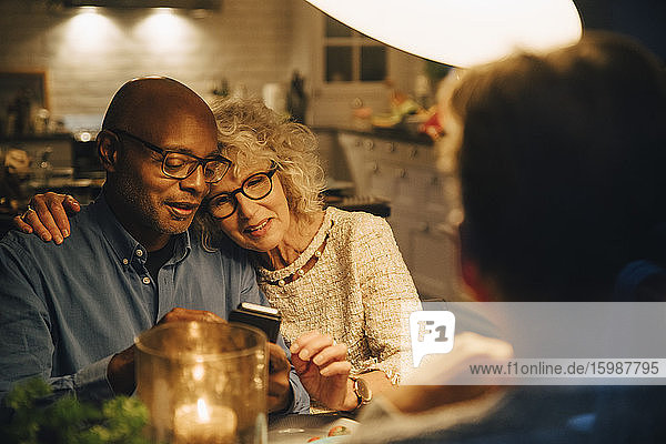 Kahlköpfiger Senior teilt Smartphone mit Frau  während er am beleuchteten Esstisch sitzt