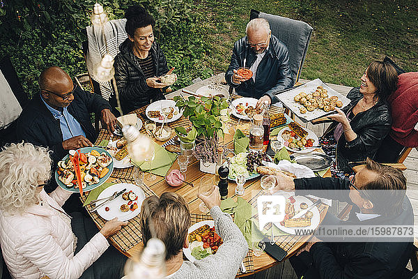 Schrägaufnahme von älteren männlichen und weiblichen Freunden  die während der Gartenparty im Hinterhof am Esstisch sitzen