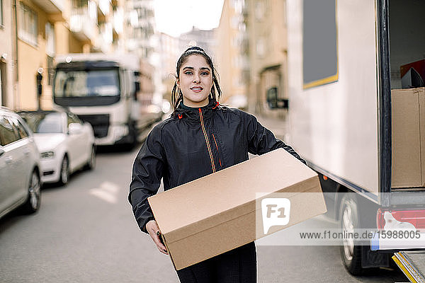 Portrait of young delivery woman carrying box while standing on street in city