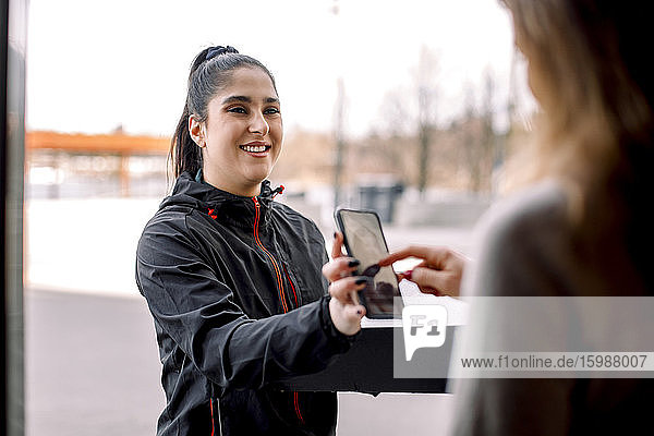 Frau unterschreibt auf Smartphone  während sie ein Paket von einer Zustellerin erhält