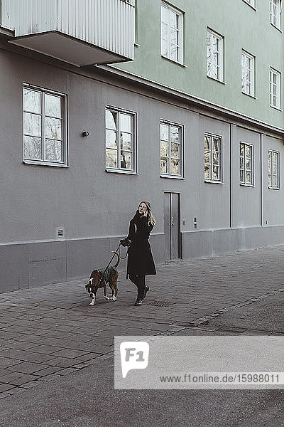 Junge Frau in voller Länge geht mit Hund auf einem Fußweg durch ein Gebäude in der Stadt