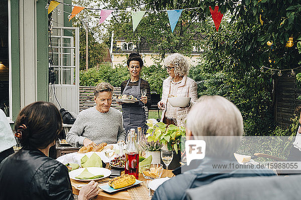 Aktive ältere Frauen bringen Freunden  die während einer Gartenparty am Esstisch sitzen  das Essen