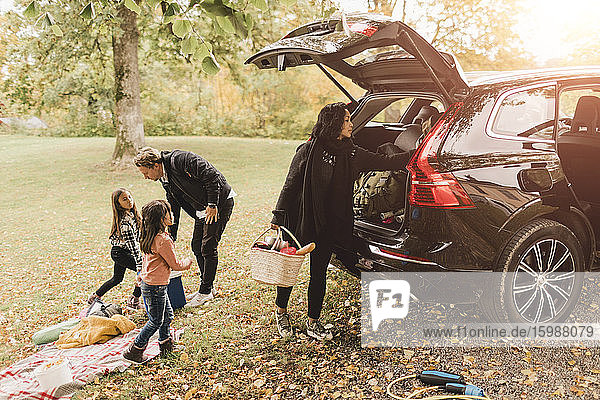 Familie entlädt Gepäck aus Elektroauto auf dem Feld während des Picknicks