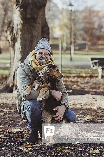 Portrait of smiling man embracing dog while crouching at park during autumn