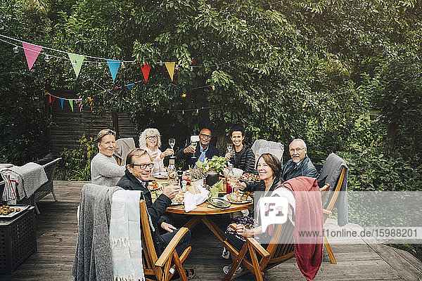 Porträt lächelnder aktiver älterer Männer und Frauen mit feierlichem Toast  die während der Party am Esstisch im Hinterhof sitzen