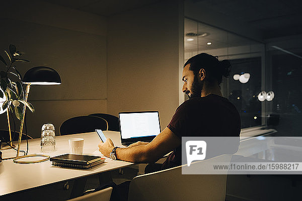 Männlicher Unternehmer  der ein Smartphone benutzt  während er mit seinem Laptop an einem beleuchteten Schreibtisch sitzt und spät im Büro arbeitet