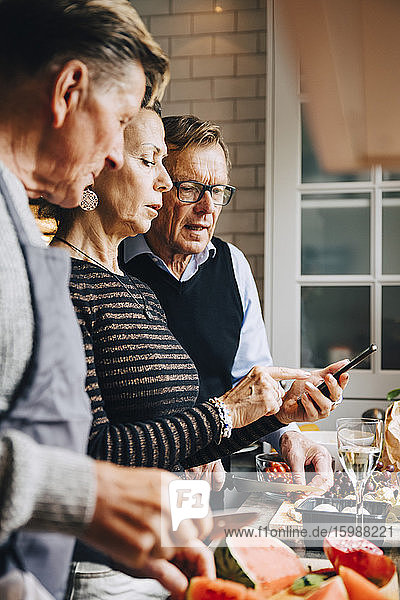 Ältere Frau diskutiert mit männlichen Freunden über Smartphone  während sie zu Hause in der Küche steht