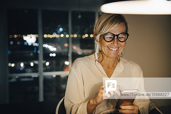Glückliche Unternehmerin nutzt Smartphone  während sie nachts im beleuchteten Büro sitzt