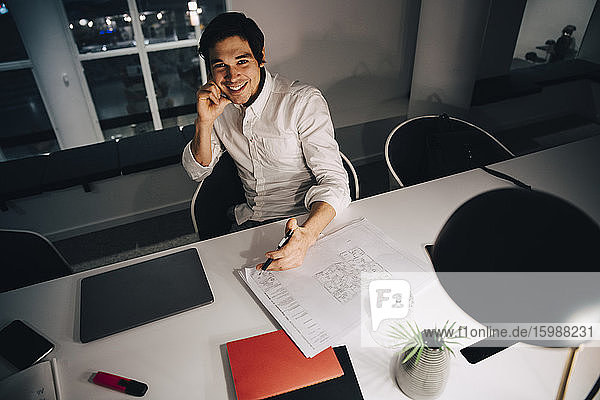Hochwinkelporträt eines lächelnden Geschäftsmannes  der mit einer Blaupause am Schreibtisch sitzt  während er spät im Büro arbeitet