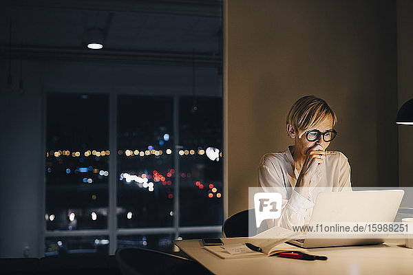 Engagierte reife Geschäftsfrau mit Laptop  während sie am beleuchteten Schreibtisch im Coworking Space sitzt