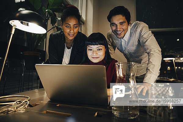 Lächelnde Geschäftskollegen planen Strategie über Laptop  während sie spät im Kreativbüro arbeiten