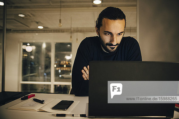 Zuversichtlicher Geschäftsmann schaut auf Laptop  während er spät im Büro arbeitet