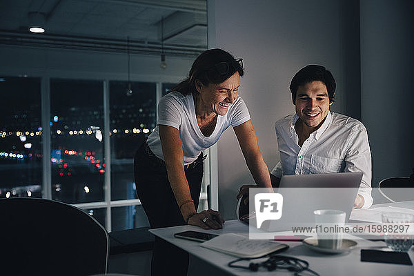 Smiling male and female colleagues discussing over laptop while working late in creative office