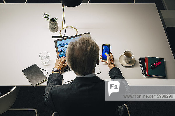 High angle rear view of senior businessman using smart phone and laptop while working late at desk in office