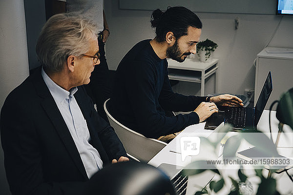 Leitender Geschäftsmann schaut während einer Sitzung am Konferenztisch im Sitzungssaal zu  während er von einem männlichen Kollegen am Laptop sitzt