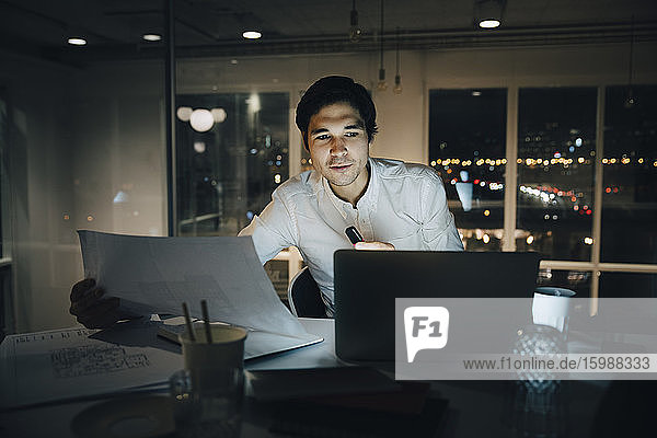 Zuversichtlicher Geschäftsmann hält Papier in der Hand  während er im Büro auf den Laptop schaut