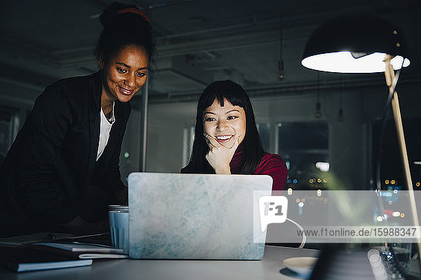 Lächelnde Unternehmerinnen diskutieren  während sie im kreativen Büro auf den Laptop schauen und lange arbeiten