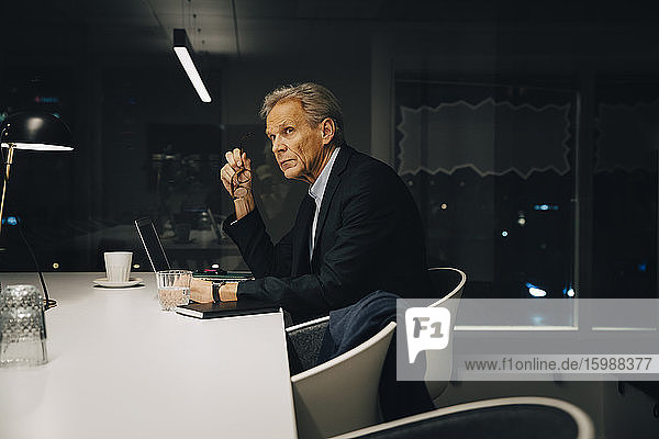 Seitenansicht eines nachdenklichen älteren männlichen Berufstätigen  der wegschaut  während er mit seinem Laptop an einem beleuchteten Schreibtisch sitzt und spät arbeitet