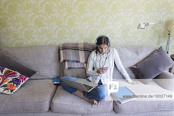 Teenagerin mit Kopfhörern und Laptop mit Smartphone auf dem Sofa