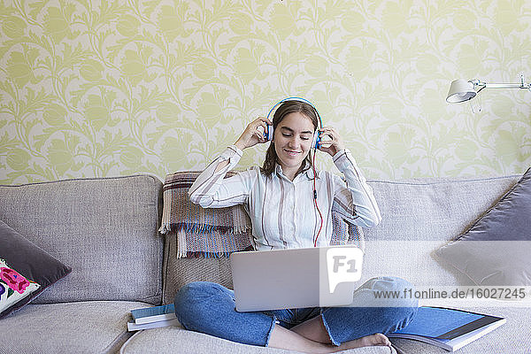 Smiling teenage girl with headphones using laptop on living room sofa