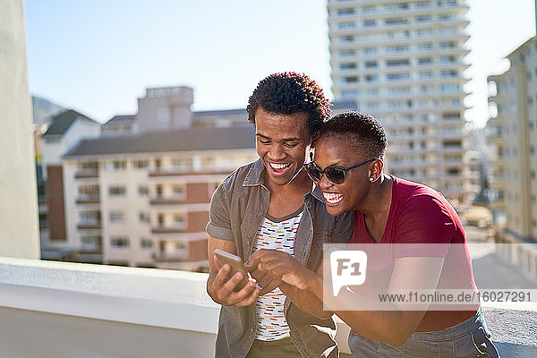 Happy young couple using smart phone on sunny urban rooftop balcony