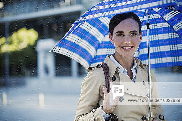 Lächelnde Geschäftsfrau im Trenchcoat unterm Regenschirm