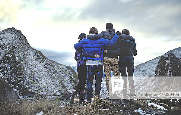 Rückansicht von vier Menschen  die Arm in Arm auf einem Berg stehen  schneebedeckte Gipfel in der Ferne.
