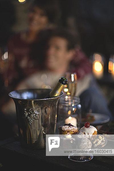 Nahaufnahme der Champagnerflasche im Weinkühler aus Metall  Kuchenstand aus Glas mit Kuchenauswahl  Person im Hintergrund.