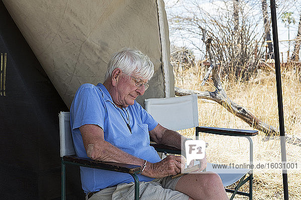 Älterer Mann  der vor einem Zelt in einem Wildsafari-Camp sitzt und ein Tagebuch schreibt.