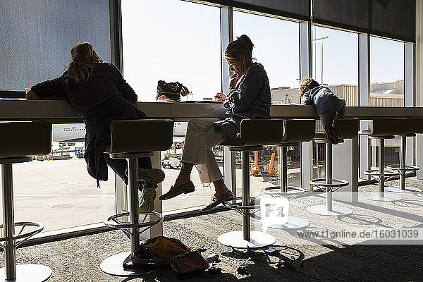 Eine reife Frau und zwei Kinder sitzen auf hohen Hockern in der Abflughalle eines Flughafens mit Blick auf das Flughafenvorfeld.