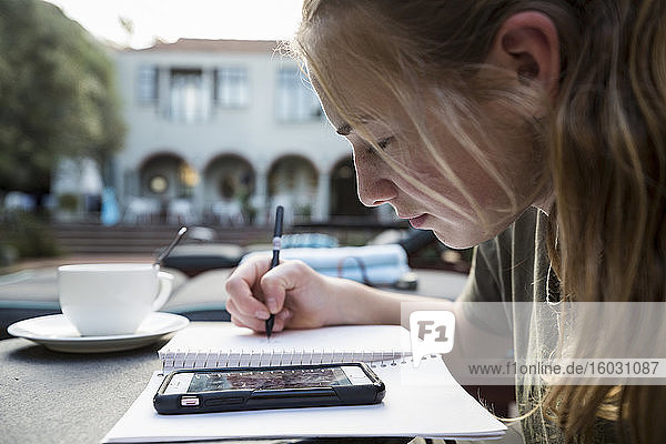 Ein Teenager  der mit Stift und Papier Notizen macht  auf ein Smartphone schaut  ein Tagebuch schreibt oder Hausaufgaben macht.