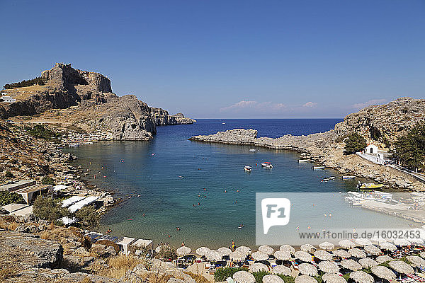 The Lindian Acropolis rises over St. Paul's Bay  a cove lined by sun shades  on a sunny day in Lindos on Rhodes  Dodecanese  Greek Islands  Greece  Europe