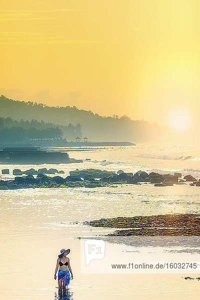 Eine junge Frau mit Sonnenhut und Schal steht in einem ruhigen Ozean  als die Morgensonne aufgeht  Bali  Indonesien  Südostasien  Asien