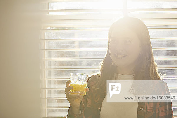 Ein junges Mädchen steht vor dem Fenster und hält ein Glas Orangensaft in der Hand.