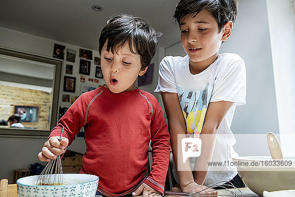 Two boys with black hair sitting at a kitchen table  baking chocolate cake.