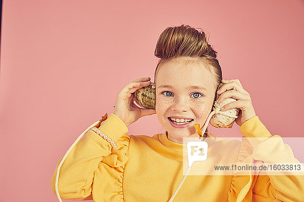 Porträt eines brünetten Mädchens in gelbem Top  das ein Muscheltelefon hält  auf rosa Hintergrund  in die Kamera blickend.