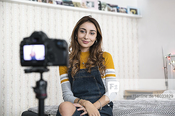 Junge Frau mit langen braunen Haaren sitzt auf dem Bett und nimmt mit einer Digitalkamera einen Blog auf.