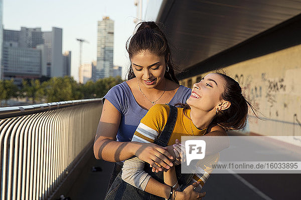 Two young women with long brown hair standing on urban bridge  hugging and smiling.