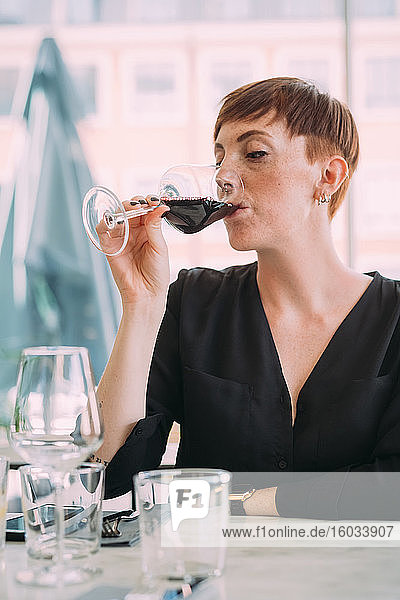 Junge Frau mit kurzen Haaren in schwarzem Top  die in einer Bar am Tisch sitzt und Rotwein trinkt.