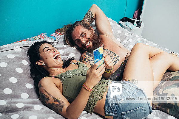 Bärtiger tätowierter Mann mit langen brünetten Haaren und Frau mit langen braunen Haaren liegen lachend auf einem Bett.