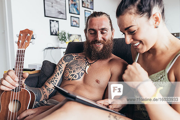 Bearded tattooed man with long brunette hair and woman with long brown hair sitting on a sofa  looking at digital tablet.